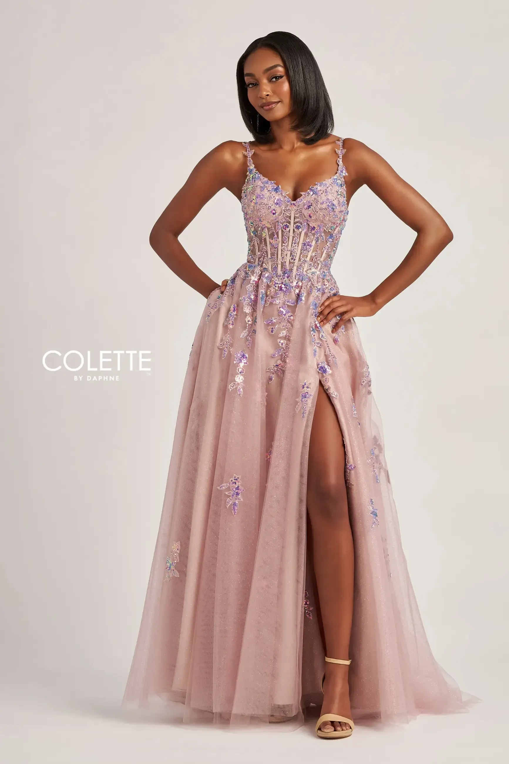 A woman in a pink gown with embellishments, posing confidently. The dress has a high slit and features floral designs.