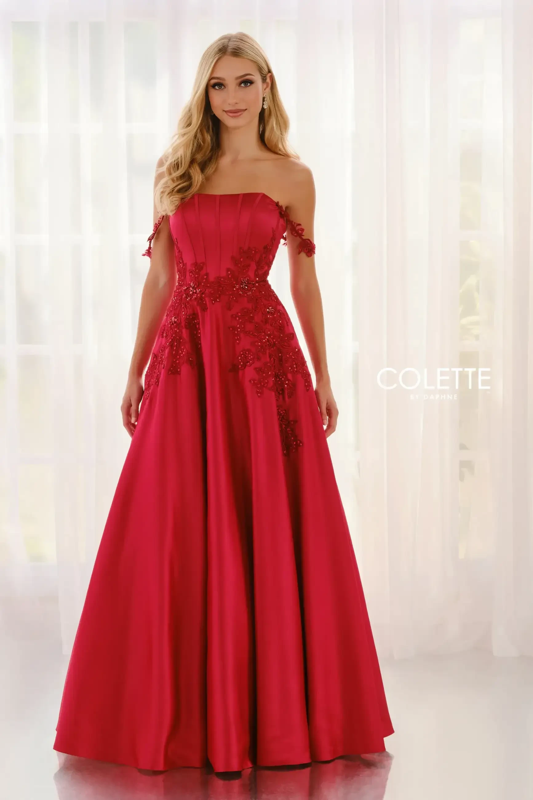 A woman stands gracefully in an elegant, off-the-shoulder red gown with intricate floral embellishments, exuding sophistication against a soft, sheer curtain backdrop.