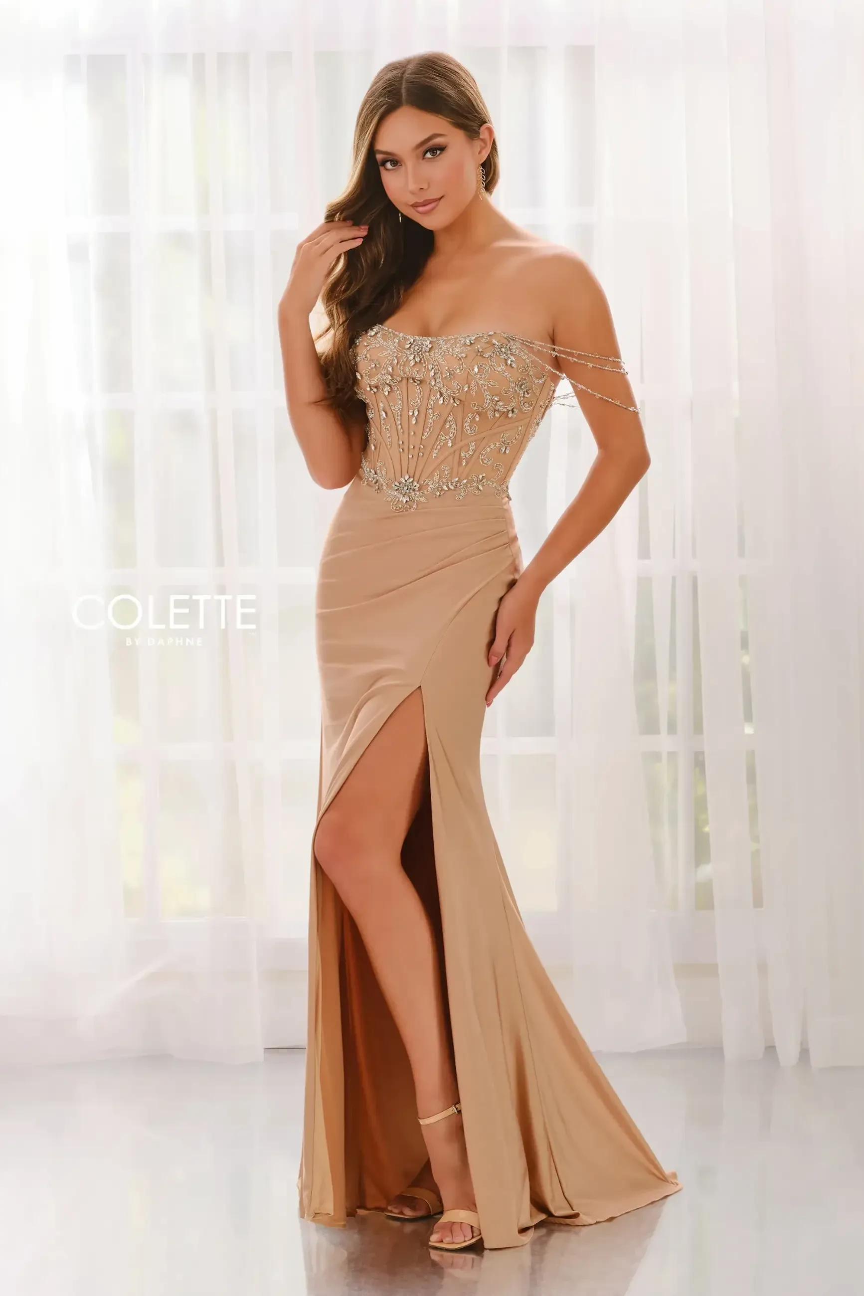 A woman in an elegant, off-shoulder champagne evening gown with intricate beadwork poses by sheer curtains. The dress has a thigh-high slit, exuding glamour.