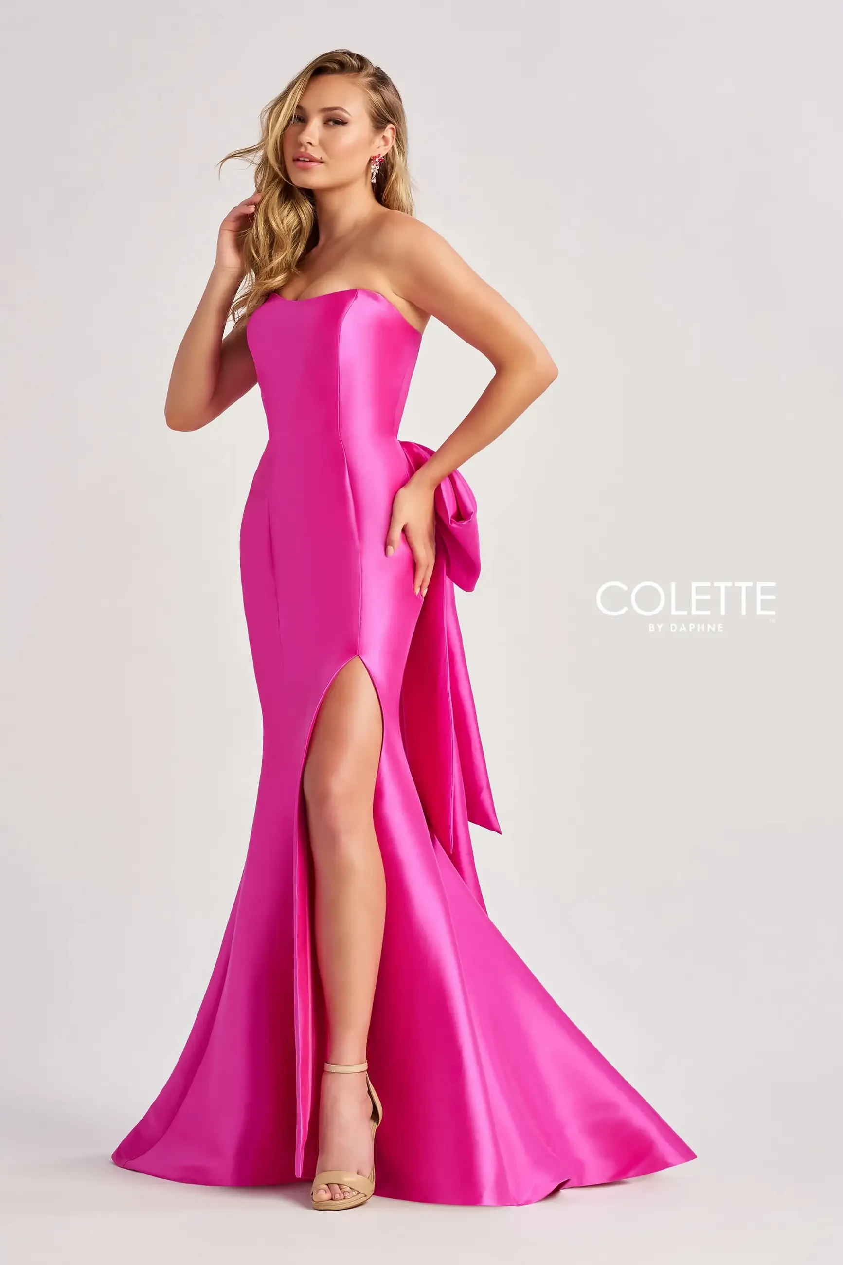 A model wearing a strapless hot pink gown with a high slit and a bow detail on the back, posing confidently.