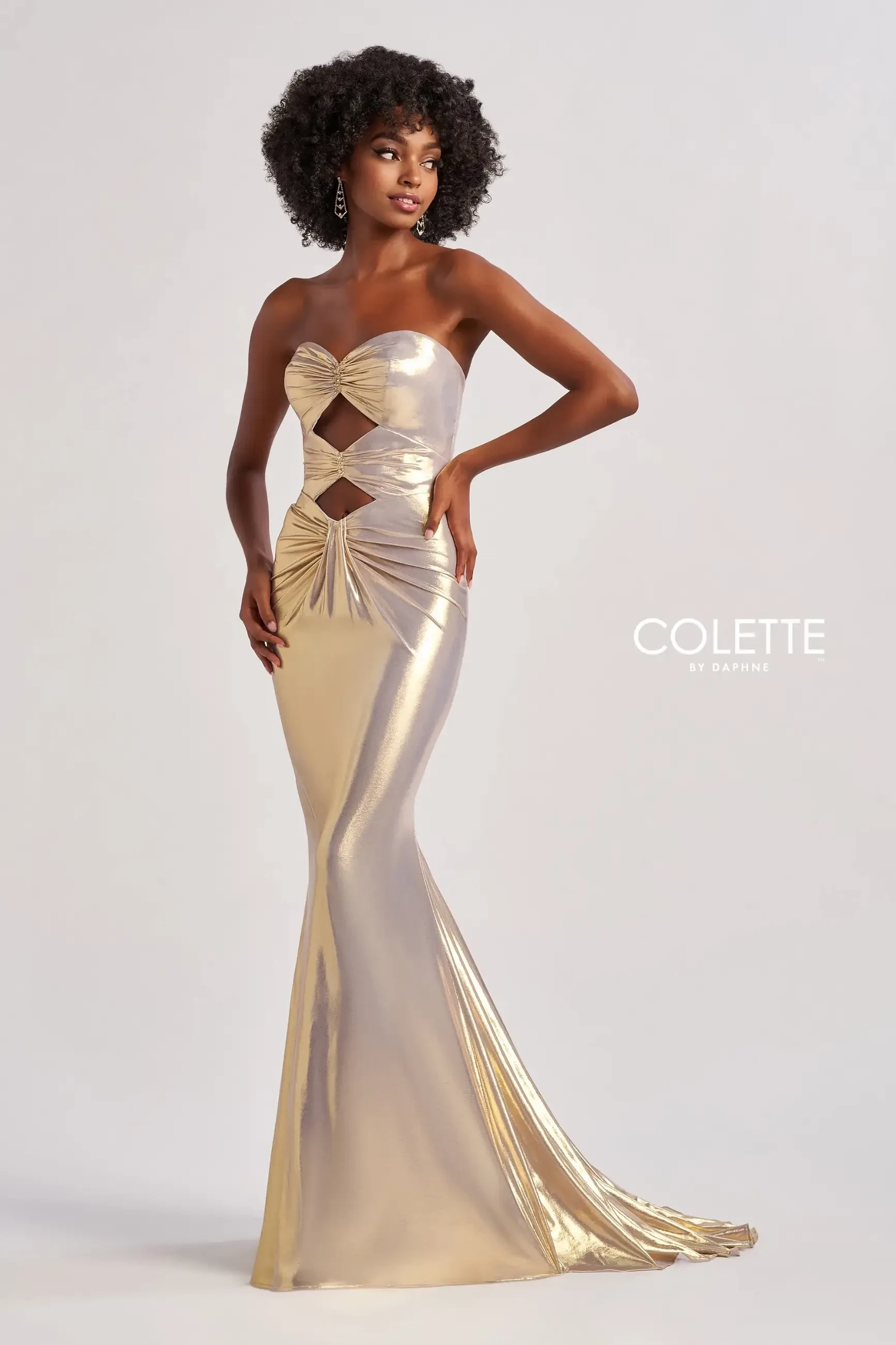 A woman in a strapless, gold metallic gown with cutouts poses confidently. The dress has a mermaid silhouette. The setting is minimalist and elegant.